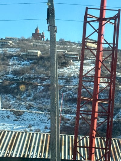 The image displays a panoramic view of a snow-dusted hillside in the city of Koghb, Armenia, under a clear blue sky. In the foreground, the top of a corrugated metal roof is visible at the bottom of the frame. Obscuring parts of the scene are a grey concrete utility pole positioned slightly left of center, extending vertically upwards, and a red metal lattice tower on the right, partially visible. Multiple thin power lines traverse the upper portion of the frame.
The midground and background depict a steep, terraced hillside partially covered with patches of snow and bare deciduous trees or shrubs, suggesting a winter or early spring season. Numerous residential structures, primarily multi-story buildings with light-colored facades and dark roofs, are scattered across the slope.
Dominating the highest point of the hill in the background is a distinctive red architectural structure, identified as the Surb Amenaprkich (Holy All-Savior) Church, which features multiple conical domes and a prominent central dome. No individuals or active interactions are observed in the scene.