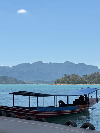 The image shows a longtail boat docked at a concrete pier. The boat has a blue awning and tires along its side to protect it from rubbing against the pier. In the background, there is a lake and mountains. The sky is blue with a small cloud. The location is Ban Ta Khun, Thailand.