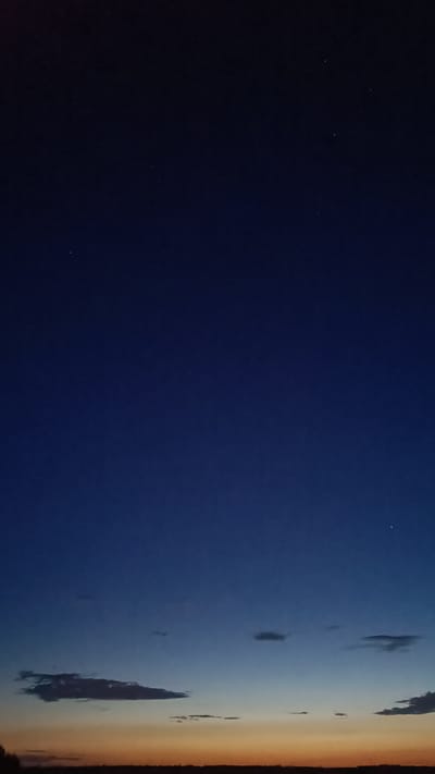 The image depicts a twilight sky over the city of Martynivs'ke, Ukraine. The sky transitions from a deep indigo at the top to a lighter blue and then to a gradient of orange and yellow near the horizon, suggesting the last moments of sunset or the first moments before sunrise. A few scattered clouds are visible, some dark and elongated, others wispy and faint. Several faint stars are visible in the darker portions of the sky. The horizon line is low and dark, indicating the presence of land, possibly fields or the outline of the city. No people or specific structures are discernible.