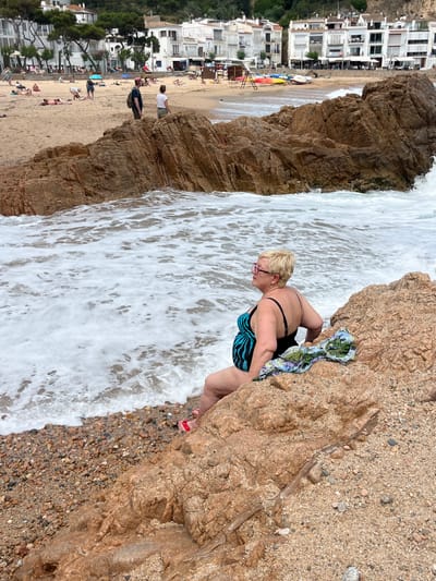 A woman in a blue and black swimsuit sits on a rocky outcropping on the beach in Tamariu, Spain. She is wearing pink sunglasses and looking to the right of the frame. The water is breaking against the rocks. The beach is sandy with small stones. The woman is wearing red sandals and there is a small blue and green sarong on the rock beside her. In the distance, there are buildings on the hillside and a sandy beach.