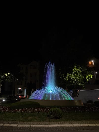The image depicts a nighttime scene in Vrnjacka Banja Municipality, Serbia, centered around a brightly illuminated fountain within a landscaped circular island. The fountain projects multiple water streams, with a central vertical jet and a cascading outer layer, all dynamically lit by internal LED lights cycling through purple, blue, and green hues. The fountain structure is encircled by a low, light-colored wall.
The island itself is planted with dark green grass, low shrubs, and beds of small red and white flowers. Surrounding the island is a paved surface featuring a cobblestone-like texture, bordered by a curb.
In the background, several multi-story buildings are partially visible through and above dense tree foliage. To the left, a building with a commercial sign displaying red text on a white background is dimly lit. A street light pole with a single lamp stands nearby. A dark-colored vehicle with illuminated headlights is visible on the road behind the island. To the right, another multi-story building shows a brightly lit upper-floor window or balcony with an orange glow. Multiple tall street light poles with downward-facing lamps are scattered throughout the background, casting light onto sections of the trees, highlighting their green leaves. Traffic signs, including a blue pedestrian crossing sign, are visible near the fountain. A white van or truck is partially visible on the road to the far right, with its red taillights illuminated. No individuals are visible in the scene.
