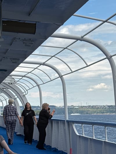 A wide shot captures the exterior walkway of a vessel sailing on a body of water, possibly the Volga River near Bet'ki, Russia. The walkway is covered by a curved, ribbed structure with a glass or transparent ceiling, offering views of a cloudy sky and a distant shoreline dotted with trees and buildings. Three women are visible on the walkway. The woman on the left wears a blue and white plaid jumpsuit and walks away from the camera. In the middle, a woman in a black t-shirt and black pants looks ahead. The woman on the right, also in black attire, is speaking on a cell phone and holding an orange object. A small white boat is visible in the distance on the water.