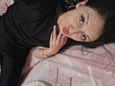 The scene depicts an indoor environment, specifically a bed, located within the city of Lanchkhuti, Georgia. One female subject is visible from the upper torso to the top of her head, lying on her back on a bed. She has dark hair, light brown eyes, dark eyebrows, and winged eyeliner. Her lips are pursed into a pout. She wears a black top, with a portion of black pants also visible, a gold-toned hoop earring in her left ear, and a gold-toned wristwatch with a rectangular face on her right wrist. Her right hand is positioned with fingers resting near her chin. The bed is covered with pink bedding featuring a repeating pattern of white, bare tree branches. The subject is angled diagonally in the frame and looking directly at the camera.
