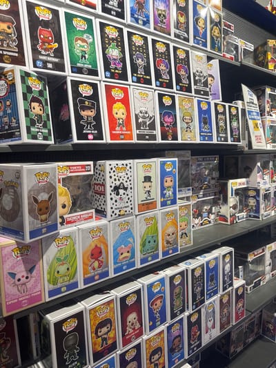 This image is a detailed, straight-on, eye-level shot of a retail display of Funko Pop! vinyl figures in their original packaging. The products are arranged on dark grey shelves, organized in rows and columns. The background is a dark grey, textured wall with some visible lighting fixtures at the top.
The Funko Pop! figures are from various franchises, including DC Comics, Marvel, Anime, and Disney. Examples include Saint Batman (580), Green Lantern (530), The Joker (606), Tanjiro Kamado (2213), Jotaro Kujo (2053), Takechi Hanagaki (2131), Eevee (577), Pikachu (884), Charmander (1157), and many more. The packaging of each figure is colorful and features an illustration of the character. Some boxes have text in English, French, and other languages. There are also some larger display boxes and some promotional materials visible on the right side of the image.
The image captures a portion of the interior of a store, likely a comic book store or a collectible shop. The specific location in Leers, France, is indicated by a sticker on one of the display boxes, which reads "LECLERC CULTURE LEERS." This suggests the scene is within a Leclerc Culture store in Leers, France. No people are visible in the image. The lighting is bright and evenly distributed, illuminating the products clearly.
