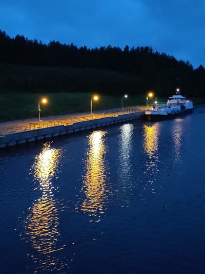 The image shows a twilight view of a canal at Devyatiny, Russia. A white barge is docked along a concrete embankment lined with a metal railing and streetlights. The streetlights cast bright yellow reflections on the dark blue water of the canal. Behind the embankment, a dense, dark green forest lines the background. The sky is a deep blue, indicating the late hour. There are no people visible in the image.