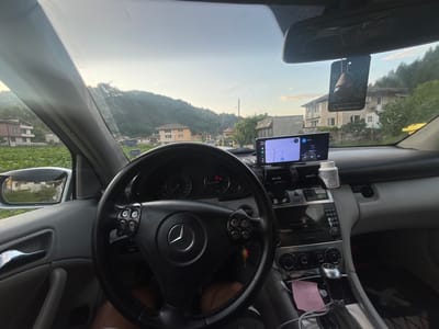 The image depicts the interior of a Mercedes-Benz W203 C-Class vehicle, observed from the driver's perspective, with an outward view through the front windshield towards a semi-rural landscape in Elhovets, Bulgaria, during the late evening.

**Vehicle Interior:**
The car features a black, three-spoke steering wheel with a central Mercedes-Benz logo. A custom aftermarket module containing four round buttons on a carbon fiber-effect panel is affixed to the right steering wheel spoke. Keys with a small keychain are visible hanging from the ignition area. The dashboard instrumentation includes a speedometer scaled to 260 km/h and a tachometer; an "SRS" warning light is illuminated. The center console houses a Mercedes-Benz audio system, climate control dials, and a metallic-finished gear shifter. A smartphone, acting as a navigation device showing a map interface, is mounted centrally on the dashboard. The phone's screen displays "20:21" and "LTE" network status. Directly below the smartphone, another smaller display indicates "Search" and "PTSD Q." A white plastic bottle with a white cap is situated on the passenger side of the dashboard. A pink sticky note with handwritten text "10:00," "04:59," and "12:30" is tucked near the gear shifter. The rearview mirror has a black, rectangular car accessory hanging from it, featuring a QR code, the text "www.BestAuto.bg," and a phone number "0887 400 004," along with a small, dark brown, pear-shaped ornament. The lower parts of the driver's legs are partially visible beneath the steering wheel, indicating a person is present in the driver's seat.

**Exterior Environment (Elhovets, Bulgaria):**
The sky is light blue with a subtle gradient, suggesting the onset of dusk or twilight. A prominent, densely forested mountain or large hill dominates the background. To the left, a two-story residential building with a light-colored facade and a brown roof is visible, positioned beyond a foreground field of low-lying green crops. To the right, several other buildings are present, including a multi-story house with balconies, multiple windows, and exterior air conditioning units, alongside a larger, more utilitarian structure. Overhead power lines and wooden utility poles traverse the scene in the middle ground. A paved road or street is visible leading away from the vehicle, with another parked car in the distance. Abundant green trees and vegetation characterize the surrounding landscape.