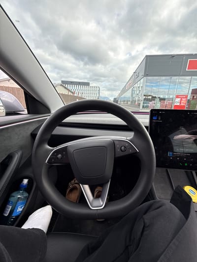 The image is taken from the interior of a Tesla, looking out through the windshield. The car appears to be parked in front of a shopping center in Harstad, Norway. The shopping center has a modern design with large glass windows and signage. The sky is overcast with dramatic clouds. Inside the car, the steering wheel and dashboard are visible. A bottle of water and a person's feet in white socks and black pants are also in the frame. The Tesla's central touchscreen displays various information, including navigation and media playback. The overall impression is that of a person inside a parked electric car, possibly waiting or taking a break.