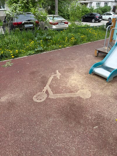 The image depicts a playground area in Tokkari, Russia. The ground is covered in reddish-brown granular surfacing, upon which a sand stencil in the shape of a scooter is visible. A metal slide is situated at the top left of the image. To the left of the playground area is a patch of grass with yellow dandelions. Partially visible parked cars are situated further to the left, beyond the grass. Building windows are visible above the parked cars. Car license plates are visible.