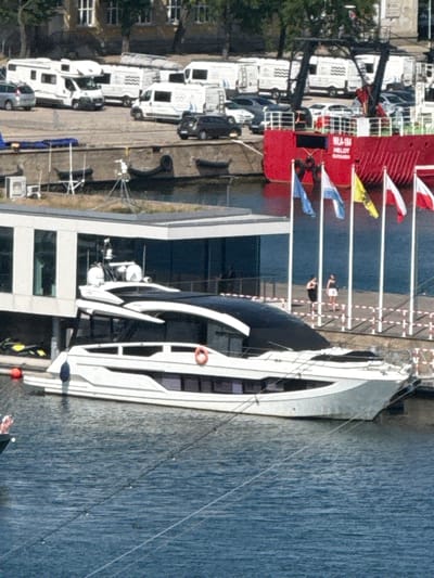 This image captures a scene at the marina in Gdynia, Poland. A large white yacht is docked in the foreground, its sleek design and dark-tinted windows visible. In the background, several white campervans are parked. A red-hulled ship with the marking "RILA-184 HELOT" is also visible, adorned with several flags. Two women stand on a walkway behind the campervans and ship. The water is calm, reflecting the blue sky and surrounding structures. The overall impression is one of a sunny day at a bustling port, with a mix of recreational and commercial vessels.