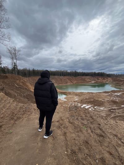 A person in a black hooded jacket and black pants stands on a dirt embankment, facing a quarry or lake. The sky is overcast. Trees line the far side of the body of water. The location is Ivlevo, Russia.