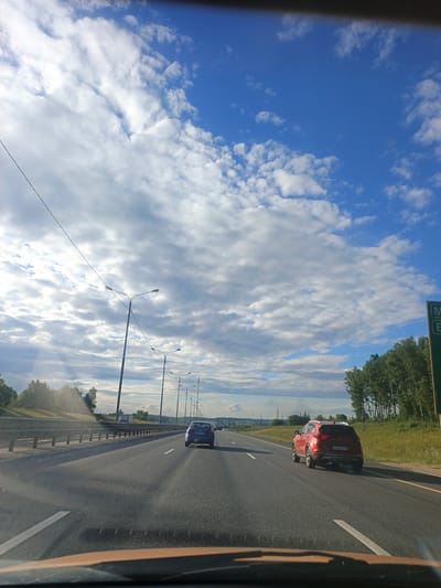 The media file shows a view from inside a vehicle traveling on a highway. The road has multiple lanes and is bordered by a guardrail on the left and a grassy area with trees on the right. There are several cars on the highway, including a blue car in the left lane and a red SUV in the right lane, both ahead of the camera's perspective. Streetlights are visible along the side of the road.

The sky is a prominent feature, with a bright blue expanse dotted with large, white, fluffy clouds. Rays of sunlight are breaking through the clouds, suggesting a partly cloudy day. A green road sign with white text is visible on the right side of the image. The time of day appears to be late afternoon or early evening, given the angle of the sun and the quality of light. The overall scene is one of travel on a suburban or rural highway. There is no discernible text that can be read clearly.