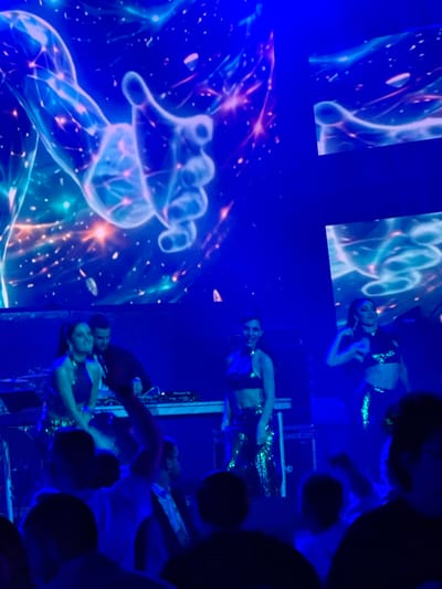 A group of people are gathered in a dimly lit venue, likely a club or concert hall, in Tsilkani, Georgia. The atmosphere is illuminated by vibrant blue and pink hues from a large screen displaying abstract, cosmic-themed visuals, possibly a nebula or starfield with glowing ethereal hands. In the foreground, on a raised platform, two women in sparkling, revealing outfits appear to be performing or dancing. One woman on the left, with dark hair pulled back, is looking down. To her right, another woman with dark hair is actively dancing, with her arm raised. Behind them, a man in a dark shirt is standing near DJ equipment. Further to the right, a third woman in a similar sparkling outfit is also dancing. The rest of the scene is filled with the silhouettes of an audience, some with their hands raised, suggesting an energetic event.