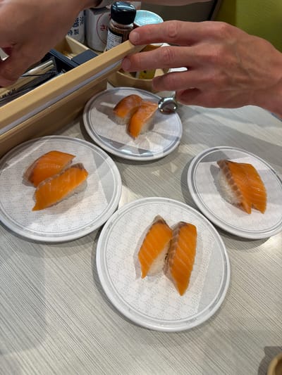 In Chiyoda, Japan, four plates of salmon nigiri sushi are arranged on a light gray, wood-grained table. The sushi is served on round, white plates with a subtle, repeating geometric pattern and a silver rim. One plate, in the upper right quadrant, holds two pieces of nigiri. To its left, another plate also contains two pieces. A third plate, in the lower right, displays two more pieces. The fourth plate, in the lower left, shows two pieces of nigiri. The hands of two individuals are visible, one on the left, opening a wooden container that holds chopsticks and packets labeled "HAM". The other hand on the right is reaching towards the sushi. In the background, there are small bottles and containers, possibly containing condiments, and a blurred green object. The lighting suggests an indoor setting.