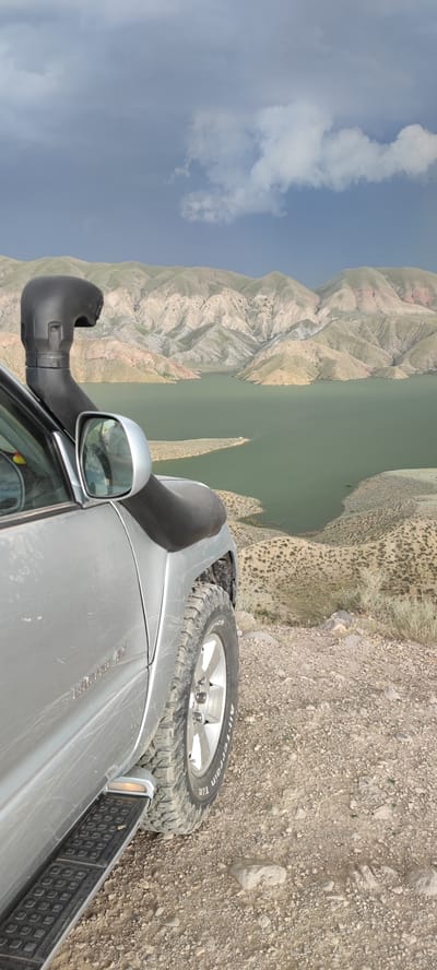 A silver SUV is parked on a rocky overlook, facing a large body of water that stretches towards a range of layered, arid mountains under a cloudy sky. The vehicle is seen from the side, with its snorkel attachment prominently displayed above the driver's side mirror. The terrain surrounding the SUV is rough and covered in loose stones and sparse vegetation. The body of water is a murky green and appears to be a reservoir. The mountains in the background exhibit a pattern of erosion, with visible valleys and peaks. The sky is a mix of dark and light clouds, suggesting potentially changeable weather. The image appears to be taken in a remote, natural landscape, possibly in the vicinity of Lanjazat, Armenia, given the geological features and the presence of a large reservoir. There are no people visible in the image.