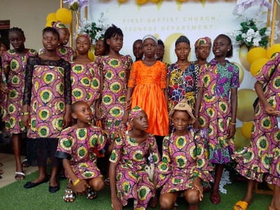 The image shows a group of young girls, possibly teenagers, dressed in colorful, patterned dresses, standing and kneeling in front of a white wall with a sign that reads "FIRST BAPTIST CHURCH, TEENAGE DEPARTMENT." The location is Yenagoa, Nigeria, based on the provided coordinates. The girls are smiling and appear to be celebrating an event or occasion. Some of the girls are wearing hats, and there are yellow balloons and other decorative elements behind them. The environment is likely a church or a community center, suggesting a religious or social context. The scene portrays a joyous and festive atmosphere.