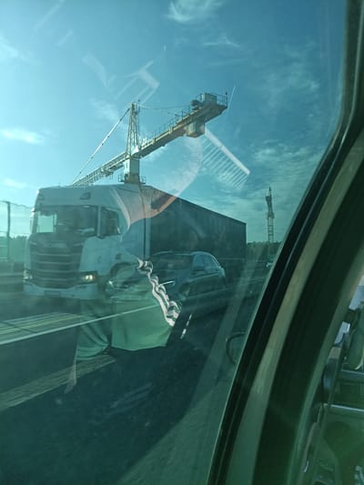 The image is a photograph taken from inside a vehicle, looking out through the windshield. The perspective is slightly angled, suggesting movement. The primary subject in the foreground is a white Scania truck, its front grille and headlights clearly visible. The truck appears to be in motion, as indicated by the blurred background and the reflection on the windshield.
Behind the truck, a large construction crane dominates the upper portion of the image. The crane is yellow and grey, with a long boom extending diagonally across the sky. A second, smaller crane is visible in the distance on the right side of the frame.
The sky is a clear blue with scattered wispy clouds. There are reflections on the windshield, most notably a reflection of a person wearing a green shirt with white stripes on the sleeves, as well as what appears to be a reflection of the interior of the vehicle, including part of the dashboard and a side mirror.
Other vehicles are present, including a dark-colored car to the right of the truck, also appearing to be in motion. The scene suggests a highway or a road under construction, given the presence of the cranes. The overall lighting indicates it is daytime, likely mid-morning or afternoon, with the sun casting light on the scene. The image captures a slice of life on a busy road with ongoing construction work in the background, viewed through the glass of a moving vehicle. The location is Hrusice, Czechia.