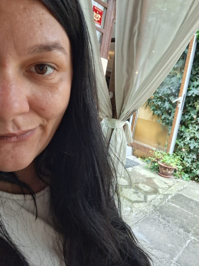 This is a close-up, rotated portrait of a woman's face and hair, taken outdoors. The woman has dark hair and brown eyes. Her face is tilted, with her nose and part of her mouth visible, as well as one eye. She appears to be looking slightly off-camera. The background suggests an outdoor setting, possibly a patio or garden area. In the lower part of the image, a stone pathway is visible, with a potted plant and dense green ivy growing around it. A sheer, light-colored curtain is draped across part of the scene, tied with a knot. Behind the curtain, a window with a yellow tint is visible, and beyond that, a wooden frame with some red signage. The lighting suggests it is daytime. The overall impression is a casual, unposed moment captured outdoors.