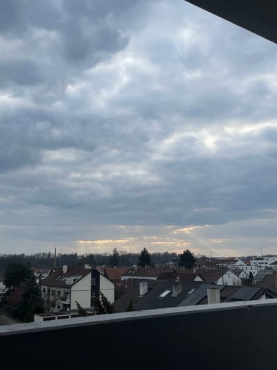 The image displays an elevated panoramic view of a residential district in Senden, Germany, observed under a predominantly cloudy sky. A dark, horizontal structure, likely a balcony railing, occupies the bottom segment of the frame. The sky is largely overcast with grey clouds, though distinct crepuscular rays of sunlight are visible penetrating the cloud cover in the mid-right and lower-central areas.
The urban landscape below consists of numerous residential buildings, characterized by dark tiled roofs, some equipped with skylights and chimneys. Multiple roofs in the mid-right section feature arrays of dark rectangular solar panels. A prominent multi-story building in the mid-left exhibits white walls with a dark vertical accent and balconies. Deciduous trees, largely devoid of foliage, are interspersed throughout the urban fabric and form a dense tree line along the distant horizon.
Above the distant tree line, a tall, slender brick chimney is visible on the left side, and a dark, pointed spire is discernible closer to the center. Two lattice-structure power line towers are present on the far right, rising above the horizon. No individuals are visible, and no specific actions or interactions are depicted within the scene.