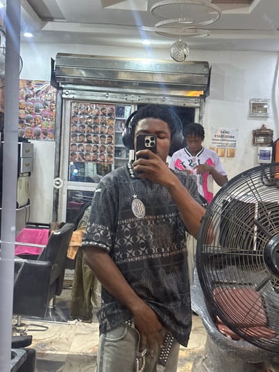The image is a selfie taken in a barber shop in Ogwashi-Uku, Nigeria, as reflected in a mirror. The photographer, a young man, is holding a phone, wearing headphones and a patterned black shirt. Behind him, a barber in a white and pink jersey is cutting the hair of another person. A wall display shows various hairstyles. Barber chairs are also visible. The foreground is partially obscured by a large fan. The overall environment suggests a typical local barber shop.