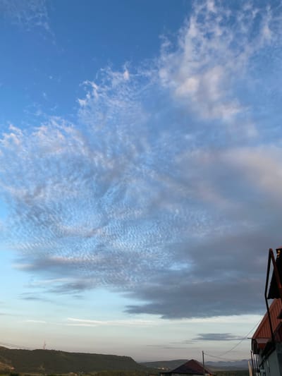 This media file captures a wide view of a sky with dramatic cloud formations above a mountainous landscape. The clouds range from thin, wispy cirrus to denser, darker formations, suggesting changing weather or atmospheric conditions. The sky transitions from a vibrant blue on the left to a lighter, hazy grey towards the right, where the sun is likely setting or has recently risen.
In the foreground, the tops of buildings with reddish-brown roofs are visible, indicating a human settlement nestled amongst the hills. One building has an overhang supported by metal poles, adding an architectural detail. The landscape features rolling hills and mountains, sparsely vegetated, with power lines and possibly a radio tower visible in the distance. The overall scene evokes a sense of natural beauty and the vastness of the sky.
The lighting suggests it is either early morning or late evening, with the sun low on the horizon. The weather appears partly cloudy, with some areas of clear blue sky and others obscured by cloud cover. There is no visible text in the image. The media file is oriented vertically, as if taken looking upwards at an angle. Given the location context of Buynaksk, Russia, the scene likely depicts a rural or semi-rural area within that region.
