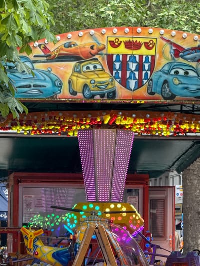 The image depicts a close-up of a colorful carnival ride, likely located in Clichy, France. The ride is adorned with cartoon car characters and illuminated by strings of red and yellow lights, as well as a panel of pink LED lights. Part of the ride's machinery and control panels are visible in the background. Trees and foliage are visible along the left side of the shot. The ride appears to be stationary, with no visible people or active motion.