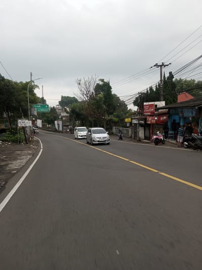The image depicts a street scene in an unidentified city, likely in Southeast Asia, characterized by a road with a yellow centerline and white edge line. On the left side of the road, there is a road sign indicating directions to "CIAWI" and "ALTERNATIF," as well as another sign with the text "BENALU TEA ASLI." A silver car is driving on the road, followed by a white van.

On the right side of the road, there is a row of small businesses or shops. People are gathered outside one of these establishments, some are standing while others are sitting. Several motorcycles are parked nearby. Overhead, there are electrical wires running along utility poles. The sky is overcast, suggesting a cloudy day.