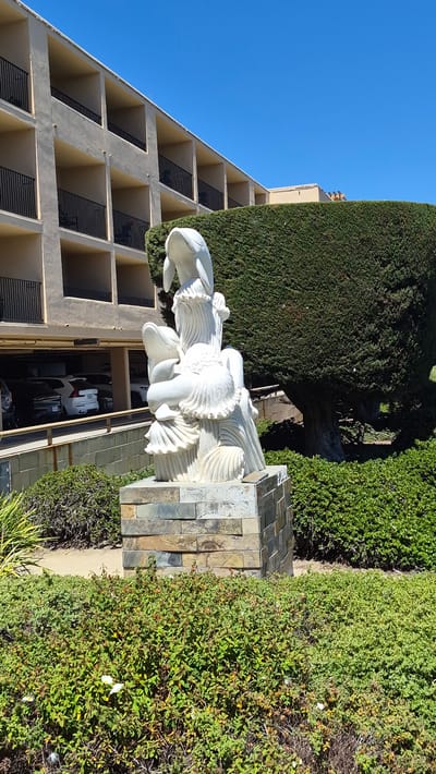 The image depicts a white sculpture of two dolphins and stylized waves, mounted on a stone base. The sculpture is situated in an outdoor setting, with a building featuring multiple levels and balconies visible in the background. A large, manicured evergreen hedge forms a backdrop to the sculpture, and a lush green bush with small white flowers is in the foreground. Several cars are parked in the open-air parking garage behind the sculpture. The sky is clear and blue. The sculpture and its surroundings suggest a coastal or resort-like environment, potentially a hotel or residential complex. The scene appears to be captured on a sunny day.