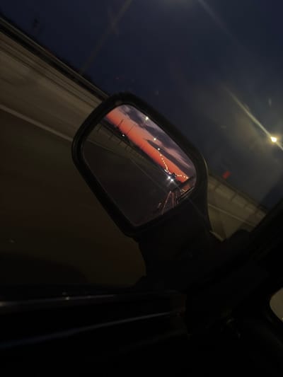 A car's side mirror reflects a highway illuminated by streetlights against a backdrop of an orange and purple sunset sky. The road ahead is dimly lit. The image suggests movement, capturing a fleeting moment during a drive at dusk. The reflection contrasts sharply with the dark interior of the car and the passing scenery on the side.