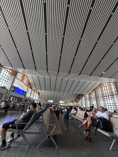 The image depicts an indoor waiting area, likely at a transportation hub in Yongding District, China. Numerous people are seated in rows of chairs, seemingly waiting. The area has a modern architectural design with a striking ceiling comprised of parallel white and black panels. A digital display shows what appears to be flight or transportation information in Chinese characters. The overall impression is that of a busy public space, possibly an airport or train station.