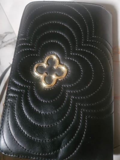 A black, quilted purse with a gold clover-shaped embellishment is the central focus of this image. The purse has a textured surface with stitched lines forming a repeating pattern of curved shapes. The gold embellishment is shiny and catches the light. The background is out of focus, but appears to be a marbled surface on the left and a textured surface on the right. The image is taken from a top-down perspective, with the purse lying flat. The location of Masaka, Nigeria is not depicted in this image.