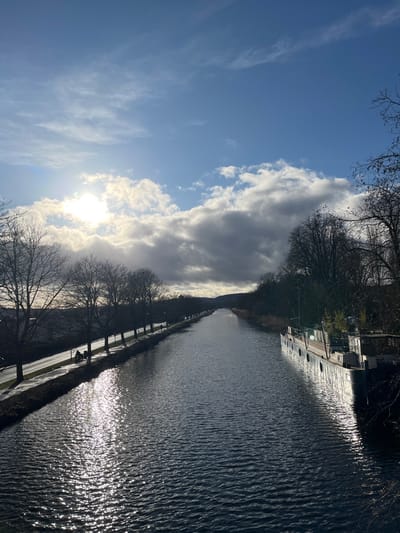 A sunlit canal view is depicted in a landscape orientation. The sky features clouds and the sun's rays, creating bright areas. The canal runs vertically with shimmering reflections. A narrow paved path bordered by trees lines the canal on the left. A building is seen at the top, behind dense trees on the opposite side of the canal. The overall tone is tranquil with contrasting light and dark elements.