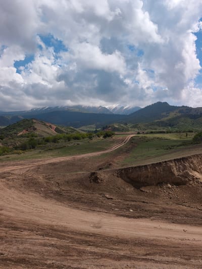 This media file presents a panoramic landscape view from Jeti-Ögüz, Kyrgyzstan, showcasing a layered natural environment under a dynamic sky. In the immediate foreground and extending into the mid-ground, a broad expanse of reddish-brown and light brown dirt is visible, possibly an area of erosion or recent earthworks. This area features wide, well-worn dirt tracks or roads that curve into the distance, suggesting pathways through the terrain. On the right side of the foreground, a steep, exposed bank or cliff of compacted earth and rock forms a natural boundary.
Moving further into the middle ground, the dirt roads lead towards a valley where patches of vibrant green grass and scattered low-lying bushes and trees begin to appear, contrasting with the bare earth of the foreground. Beyond these green patches, undulating hills, some covered with dense coniferous forests, rise gracefully. In the distant background, majestic mountain ranges with striking snow-capped peaks stretch across the horizon, partially veiled by the cloud cover.
The sky is a prominent feature, dominated by large, billowy cumulus clouds in shades of white and grey, interspersed with patches of clear blue sky, indicating a partly cloudy day. The lighting suggests it is daytime, likely late morning or early afternoon, with diffused light illuminating the landscape and casting subtle shadows. There are no people, animals, or visible text in the image. The scene conveys a sense of vastness, rugged natural beauty, and tranquility characteristic of mountainous regions.