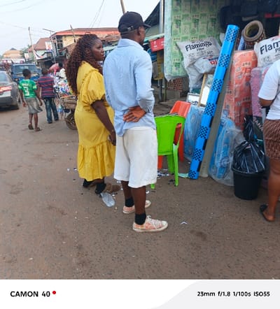A medium shot depicts an outdoor street scene with people near a roadside shop. A woman in a yellow dress stands next to a man in a blue shirt and white shorts. Other people are walking behind them. The shop displays items like pillows and blue PVC pipes. A green chair and a black bucket are also visible. The street is paved, with litter scattered around. The photo includes camera details at the bottom: "CAMON 40" and "23mm f/1.8 1/100s ISO55".
