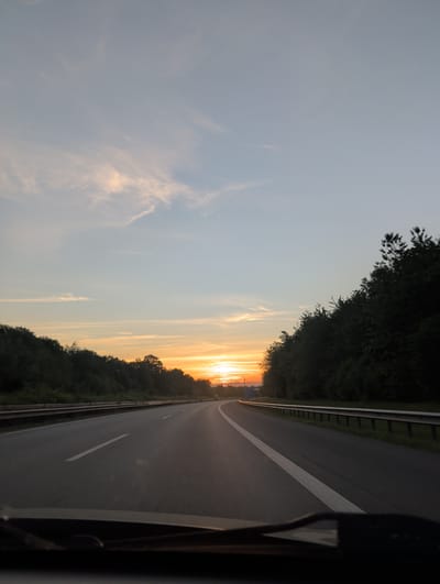 The image depicts a highway at sunset, looking forward from the perspective of a vehicle's dashboard. The sky is a gradient of pale blue and orange, with wispy clouds catching the last rays of the sun. The highway itself is asphalt, with clear white lane markings. On either side of the road, there are dense lines of trees, silhouetted against the bright sunset. A metal guardrail runs along the right side of the highway. The scene is serene and appears to be on a rural highway, possibly on the outskirts of Polch, Germany, given the information provided. No people are visible.