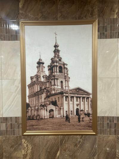 The image shows a framed sepia-toned photograph of the Church of the Assumption of the Blessed Virgin Mary in Kursk, Russia. The church is a grand baroque structure with multiple domes and a tall bell tower topped with a cross. In the foreground, several people in historical attire are visible, along with horse-drawn carriages and a horse. The photograph appears to be old, possibly from the late 19th or early 20th century, judging by the style of the clothing and the presence of horse-drawn transport. The church is depicted in a somewhat elevated perspective, with its facade facing the viewer. The surrounding environment is a paved area, likely a square or street in front of the church. The photograph is framed with a wide, ornate golden frame.
