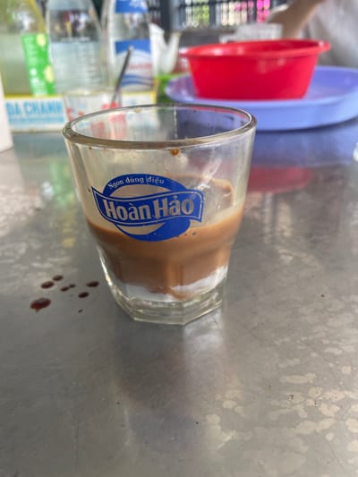 The scene shows a partially consumed beverage in a glass sitting on a metallic surface, likely a table or counter. The glass has a blue logo with white text on it, possibly a brand name. Brown droplets or stains are present around the glass. In the blurred background, there are colorful plastic bowls stacked and what appear to be bottles with labels in Vietnamese. The location is Nam Ninh Hoa, Vietnam. There is no information about people, actions, or interactions. The environment is characterized by a utilitarian and casual setting, potentially a street food stall or a simple eatery.