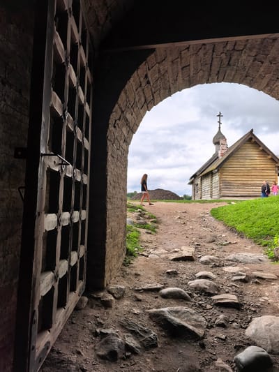 The image captures a view through a massive, weathered wooden gate and stone archway in Staraya Ladoga, Russia. The gate, constructed of thick wooden planks and metal fixtures, is partially open, revealing a rocky, dirt path leading towards a small, wooden church with a domed roof and a cross. The church is situated on a grassy knoll under a cloudy sky. A young woman with long, dark hair, wearing a black t-shirt and white shorts, walks away from the viewer on the path. Further down the path, near the church, two other figures, possibly children, are visible. The ground in the foreground is uneven, covered with large stones and loose dirt, suggesting an ancient or historical site. The stonework of the archway and the gate indicates a fortress or a similar historical structure.