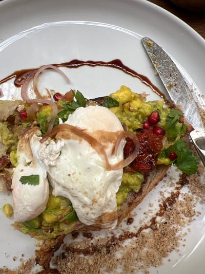 On a white plate, is a slice of toast topped with avocado, cilantro, pomegranate seeds, a roasted tomato, and a poached egg, drizzled with balsamic glaze and a sprinkle of what appears to be a nut crumble. A knife rests at the top left of the plate. The location is Rumassala, Sri Lanka.
