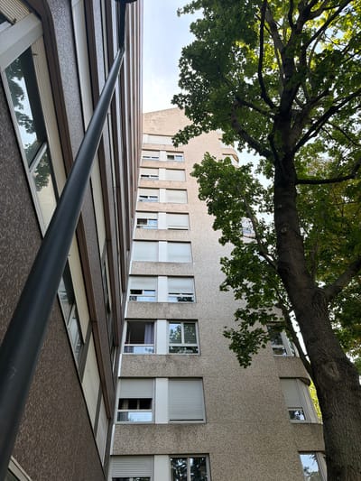 In Courbevoie, France, two apartment buildings are viewed from a low angle, looking upwards. On the left, a darker, brick-clad building features a series of rectangular windows, some angled. A tall, dark grey pole runs vertically along the left side of the frame, partially obscuring the building. To the right of this building, a lighter-colored apartment building with a textured facade rises higher. This building has numerous rows of windows, many covered with horizontal blinds. A large, leafy tree with a thick trunk is positioned in the foreground on the right side of the frame, its branches and green foliage extending across the upper portion of the image and partially obscuring the sky and parts of the lighter building. The sky above is clear and light blue. The scene appears to be an outdoor urban environment during daylight.