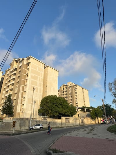 In Perímetro Urbano Barranquilla, Colombia, a low-angle shot captures two tall, light-colored apartment buildings against a bright blue sky dotted with scattered white clouds. Power lines stretch across the top of the frame. In the foreground, a paved road curves to the left, with a brick-patterned sidewalk on the right. A silver car is parked near a low concrete wall, which features decorative grates. A woman is crossing the street, carrying a red object, and walking towards the right side of the image. A street light stands near the parked car. Trees with lush green foliage are visible between the buildings and along the wall. The buildings appear to be under construction or renovation, with some sections of their facades showing unfinished concrete. The lighting suggests it is late afternoon.