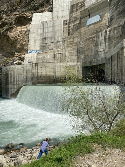 The image depicts the Chirkeyskaya Hydroelectric Power Station, located in the Untsukulsky District of Dagestan, Russia, near the village of Irganay. A vast concrete dam dominates the scene, with water cascading over its crest, creating a powerful waterfall. The water is a turbulent, greenish-blue, rushing into the river below. On the rocky bank in the foreground, a person wearing a blue shirt and pants is bent over, appearing to examine the rocks. Green vegetation grows along the bank, and a few trees with bare branches are present, adding to the rugged, natural setting. The concrete structure of the dam is marked with graffiti, with some illegible writing and symbols visible. The sky is clear and bright, indicating a sunny day.