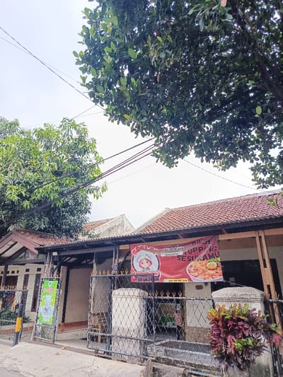 A restaurant with a red banner hanging in front of the building with the words "Mie Ayam Topping Sesukamu". A cartoon character is on the left side of the banner. The restaurant is behind a black gate with ornate designs. Trees overhang the restaurant. A green poster is visible on the left side of the gate. The sky is overcast.