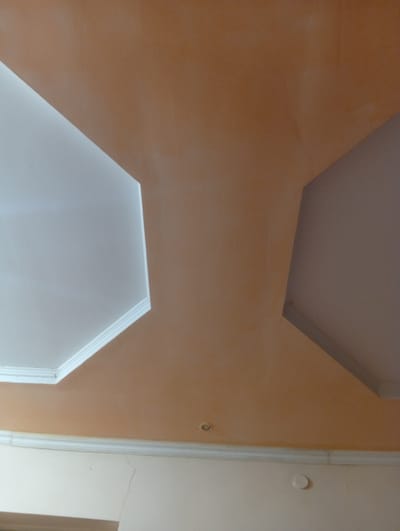 The image shows a close-up view of a ceiling with a beige upper section and a white lower section. The beige section features two recessed, hexagonal ceiling designs, one on the left and one on the right, both illuminated from within. A white decorative molding runs along the junction between the ceiling and the wall, which is painted a lighter shade of beige. A visible crack runs through the wall in the lower left portion of the image, near the molding. There are two small, circular fixtures visible on the wall in the lower half of the image, one to the left and one to the right of the crack. The image appears to have been taken indoors, likely in a residential or commercial space. The specific location in Жартытөбе ауылдық округі, Kazakhstan, is not discernible from the visual elements present in the photograph.