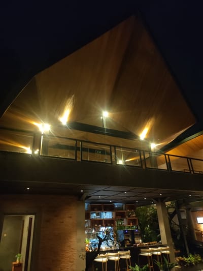 The image depicts the exterior of a modern, multi-level building at night, located in Mas, Indonesia. The upper section features a distinctive pitched roof with light-brown wood paneling illuminated by multiple warm-toned lights, creating starburst lens flare effects. A second-story balcony or mezzanine level is visible, enclosed by clear glass panels with black frames and a dark railing. The lower section of the building includes a brick-clad wall on the left, adjacent to an open doorway revealing a dimly lit interior. To the right, a ground-floor bar area is present, featuring a dark counter and several light-colored bar stools with metallic bases. Behind the bar, a wooden shelving unit displays bottles and glasses. Green foliage is visible around the bar, and a dark, stylized tree-like object stands on the counter. The overall environment is characterized by contemporary architecture and artificial illumination against a dark night sky. No individuals are visible.