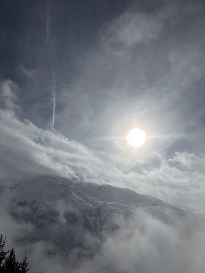 The image displays a snow-covered mountain landscape in the city of Andermatt, Switzerland, under a partly cloudy sky. An intense, bright sun with significant lens flare and glare is situated in the upper right quadrant. A distinct, white linear contrail extends vertically through the upper-left sky, crossing a patch of clear blue-gray sky before entering a cloudy area. Snow-dusted mountain peaks and slopes dominate the mid-ground and background, partially obscured by dense clouds and mist that rise from the valleys and enshroud the lower portions of the mountains. Dark, coniferous tree tops are visible in the bottom-left foreground, emerging from the atmospheric conditions. No human subjects, specific actions, or man-made structures are identifiable within the frame.