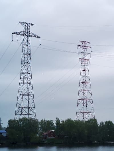 The image shows two high-voltage power line support towers located in Konakovo, Russia. The tower on the left is painted in dark grey and the tower on the right is painted in white and red. The towers are situated on a strip of land next to a body of water. On the shore, behind the strip of land, there are some buildings and trees. The sky is overcast and grey. There are thin power lines running from one tower to the other.
