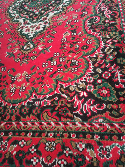 A close-up, slightly angled shot shows a red rug with a complex, traditional pattern. The pattern features interwoven floral and paisley motifs in black, white, green, and brown. The texture of the rug appears somewhat worn, with individual threads visible. The background is entirely filled with the rug's surface, providing no context of its surroundings or location within Syrdarya, Uzbekistan.