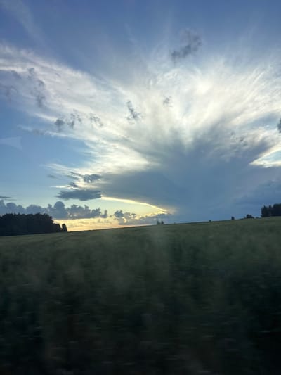 The image shows a landscape scene with a dramatic sky and a field. The sky is filled with a mixture of white and gray clouds, with a bright light breaking through the clouds on the right side of the frame. The clouds are wispy and feathery in some areas, and more dense and turbulent in others. The horizon is marked by a line of dark green trees on the left, and scattered trees on the right. Between the sky and the treeline is a rolling green field, which appears to be a meadow or pasture. The field is mostly dark green, with some lighter patches. The overall impression is one of a rural landscape, possibly at dusk or dawn, given the lighting. The image does not contain any identifiable landmarks or structures that would suggest a specific location within the city of Пермяки, Russia.