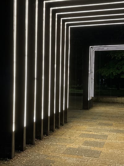 The image depicts illuminated steps in Vrnjacka Banja Municipality, Serbia. The steps are dark and have bright white lights running along the edges of each step, creating a linear pattern. The steps lead from a dark area with trees towards a paved area with rectangular stones. The overall atmosphere is nocturnal and modern.