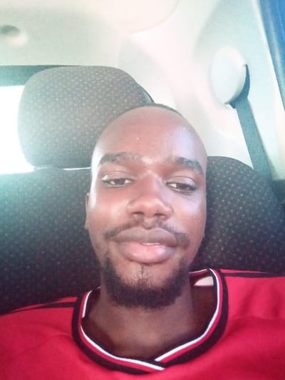 A headshot-style image depicts a single male individual with dark skin, a short beard, and short dark hair, situated within the interior of a vehicle. The individual is looking directly at the camera with a neutral to slight smirk expression. He is wearing a red collared shirt with black and white stripes along the collar edge. Behind him, the interior of a vehicle is visible, including dark grey, textured headrests, a light-colored roof lining, and a black seatbelt on the right. Natural light appears to illuminate the scene, primarily from the left. The scene is described as being in Majengo ward, Kenya.